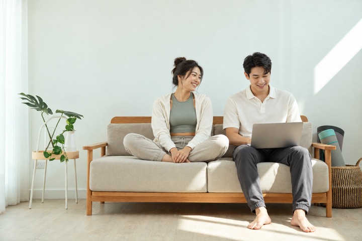 Ảnh minh họa AI: một người phụ nữ ngồi xếp bằng trên sofa, mỉm cười nhìn người đàn ông bên cạnh đang dùng laptop, trong căn phòng sáng có cây xanh.